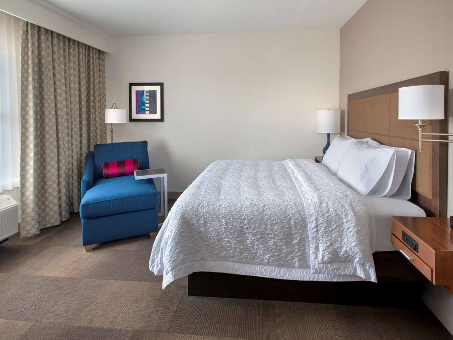 A hotel room with a large bed, a blue armchair, a small table, and a wall-mounted lamp.