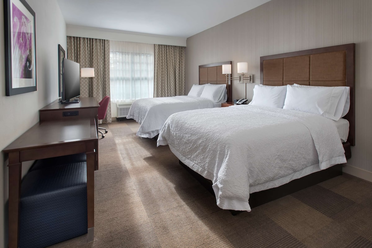 A hotel room with two beds, a desk, a chair, a TV, and a window with curtains.