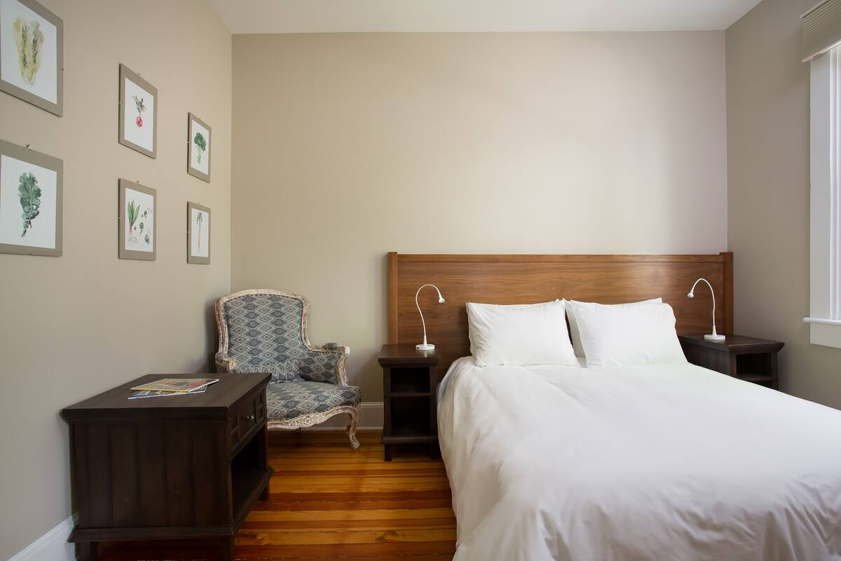 A bedroom with a wooden headboard, a single bed, a nightstand with a lamp, a chair, and framed botanical prints on the wall.