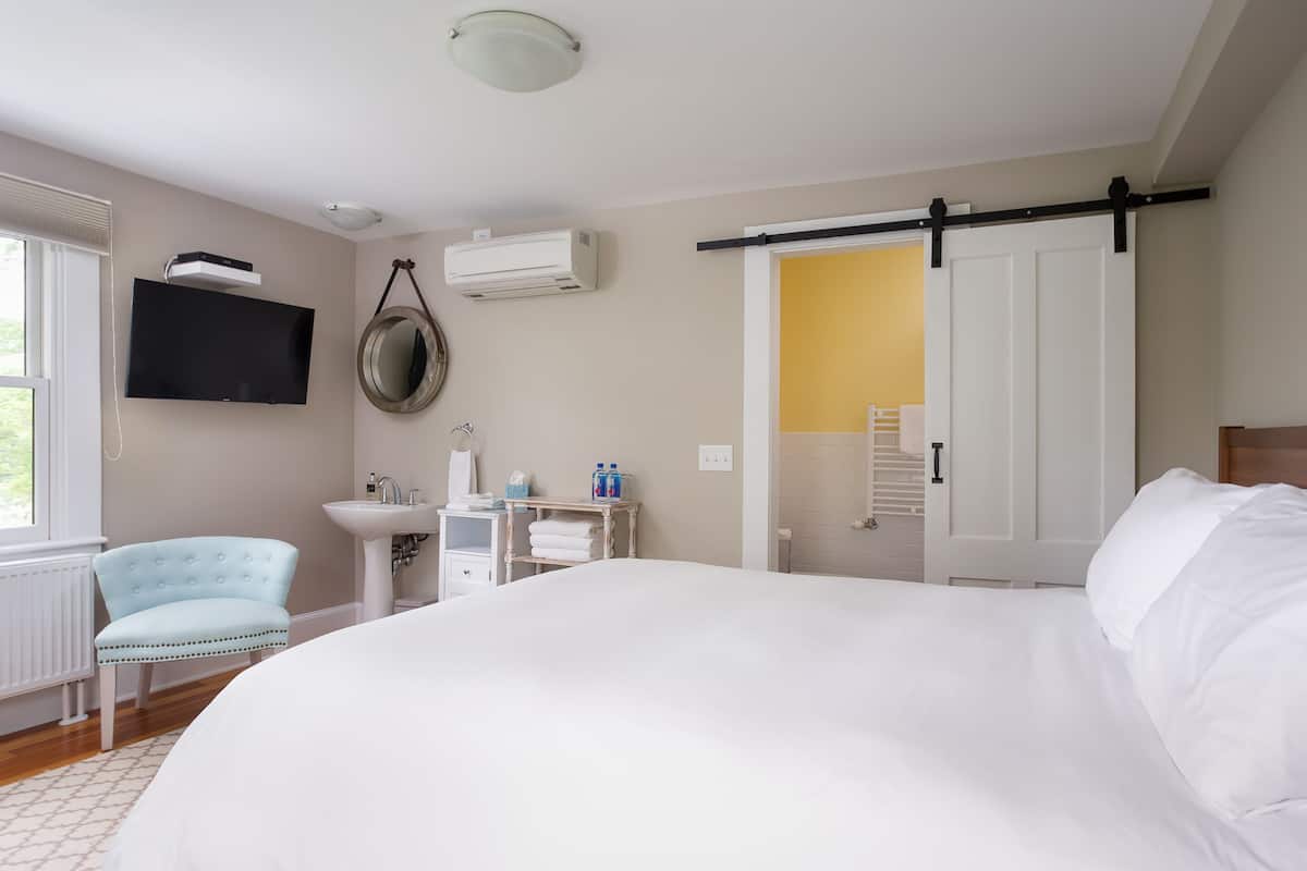 A hotel room with a large bed, a flat-screen TV mounted on the wall, a bedside table with a mirror, a sink, and a sliding door leading to a bathroom.