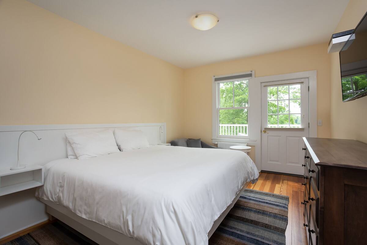 A bedroom with a bed, bedside table, dresser, and a window with a view of trees.