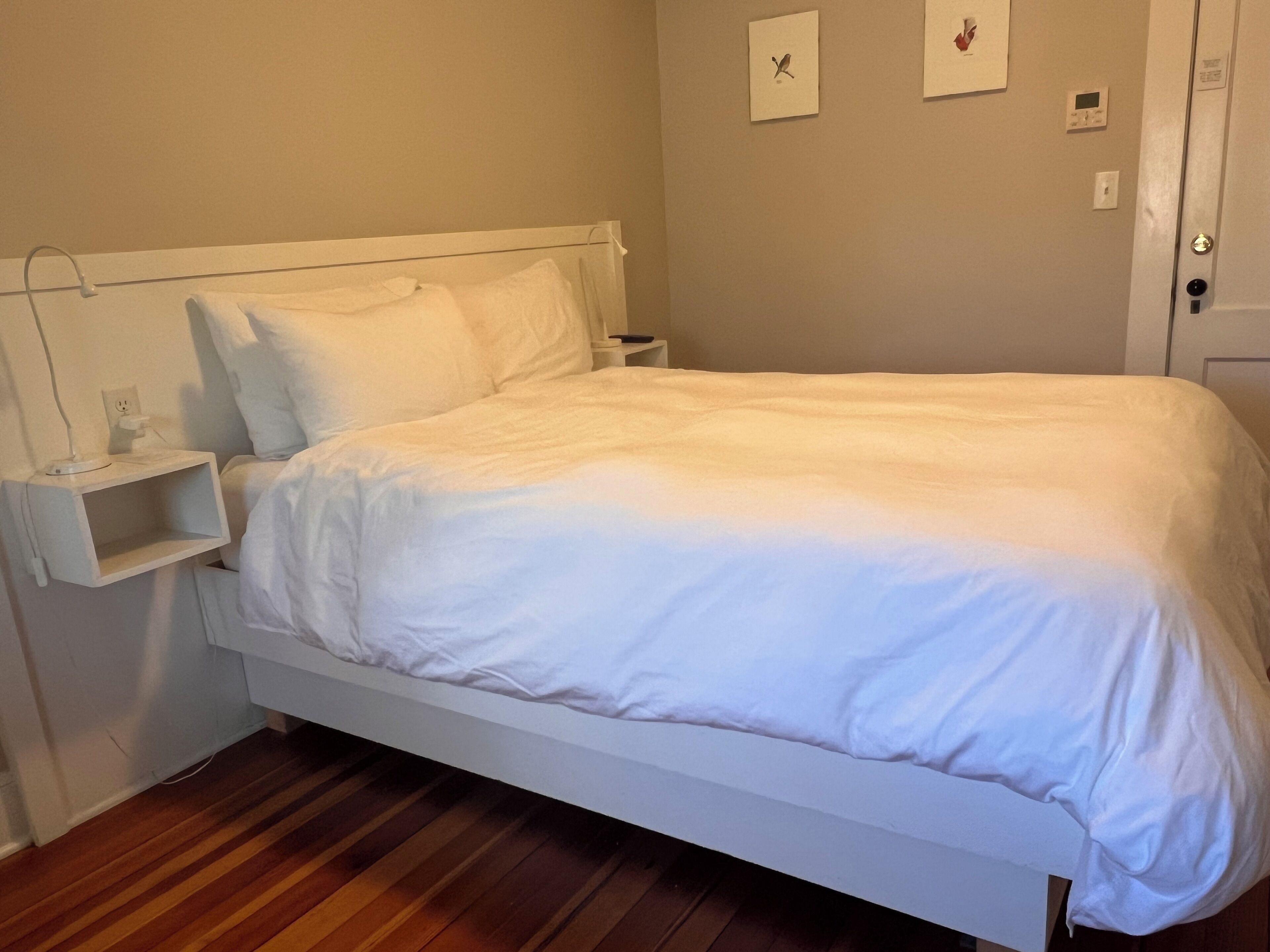A neatly made bed in a room with a wooden floor, a bedside table with a lamp, and a wall-mounted thermostat.