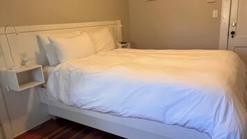 A neatly made bed in a room with a wooden floor, a bedside table with a lamp, and a wall-mounted thermostat.