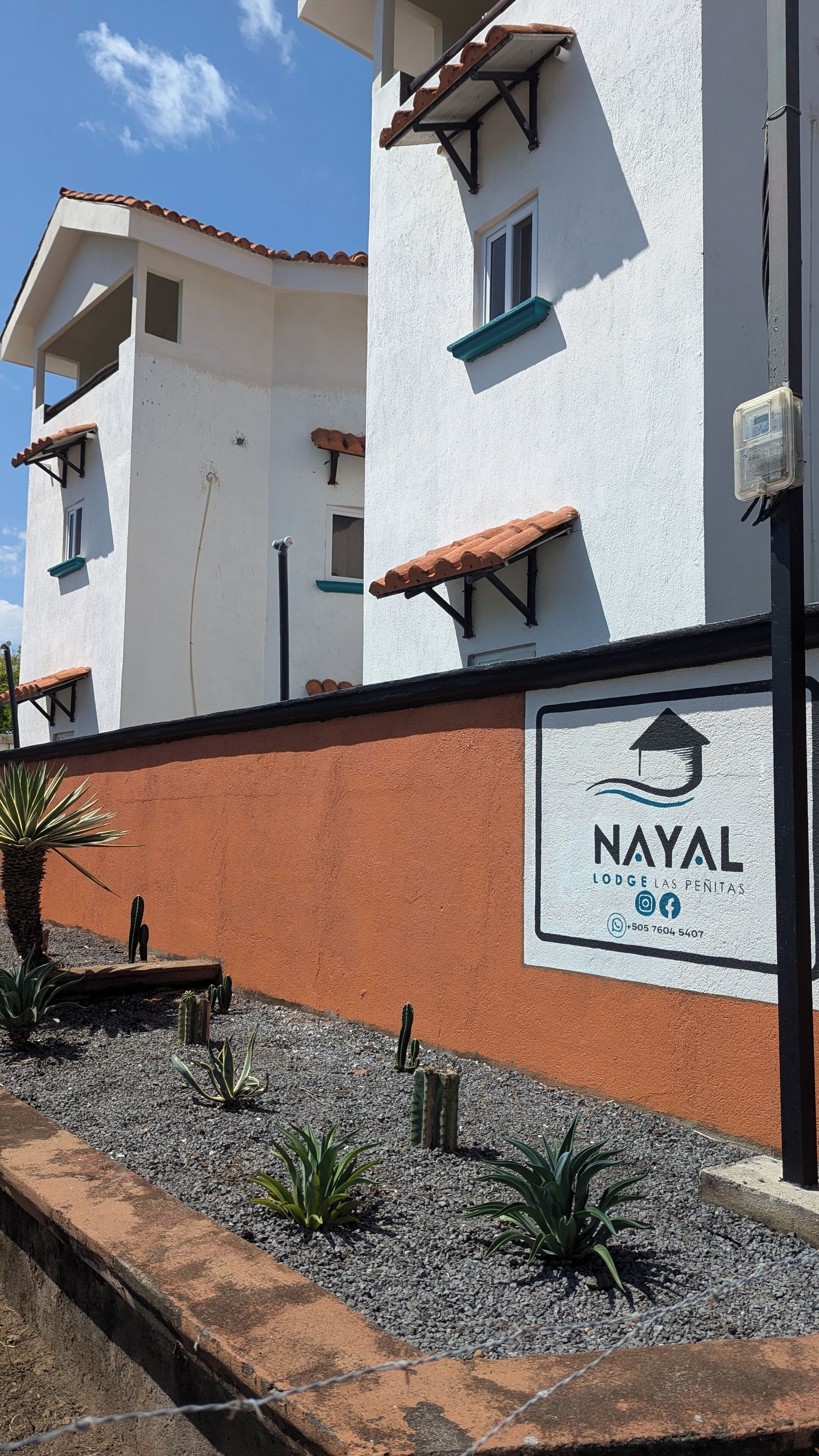 Um edifício com fachada branca, parede vermelha e placa com a inscrição 'NAYAL LODGE LAS PENITAS' e informações de contato.