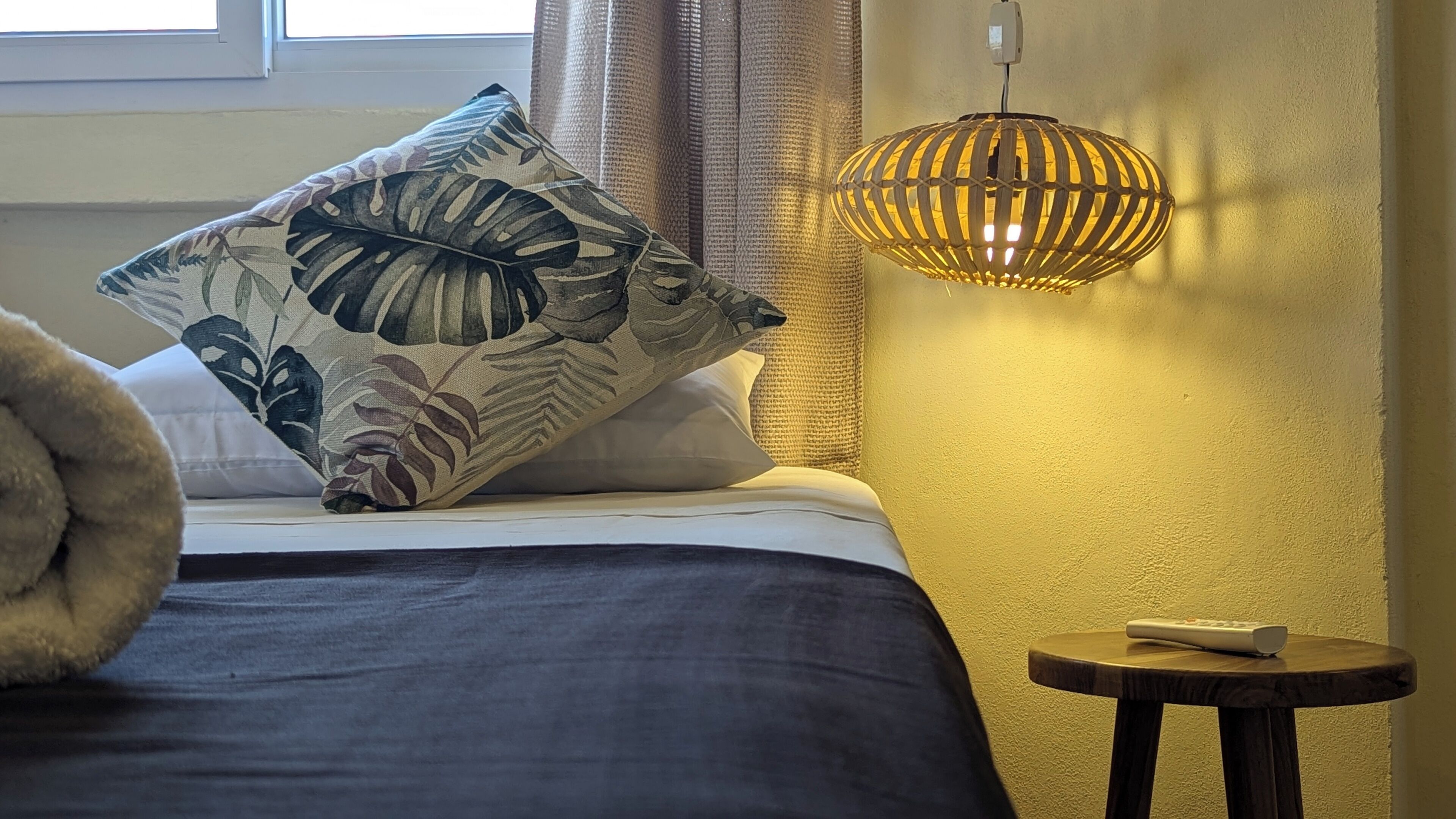 A bed with a large leaf-patterned pillow, a bedside table with a remote control, and a hanging lamp.