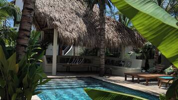 Zona de piscina con una estructura de techo de paja, sillas de descanso y palmeras.