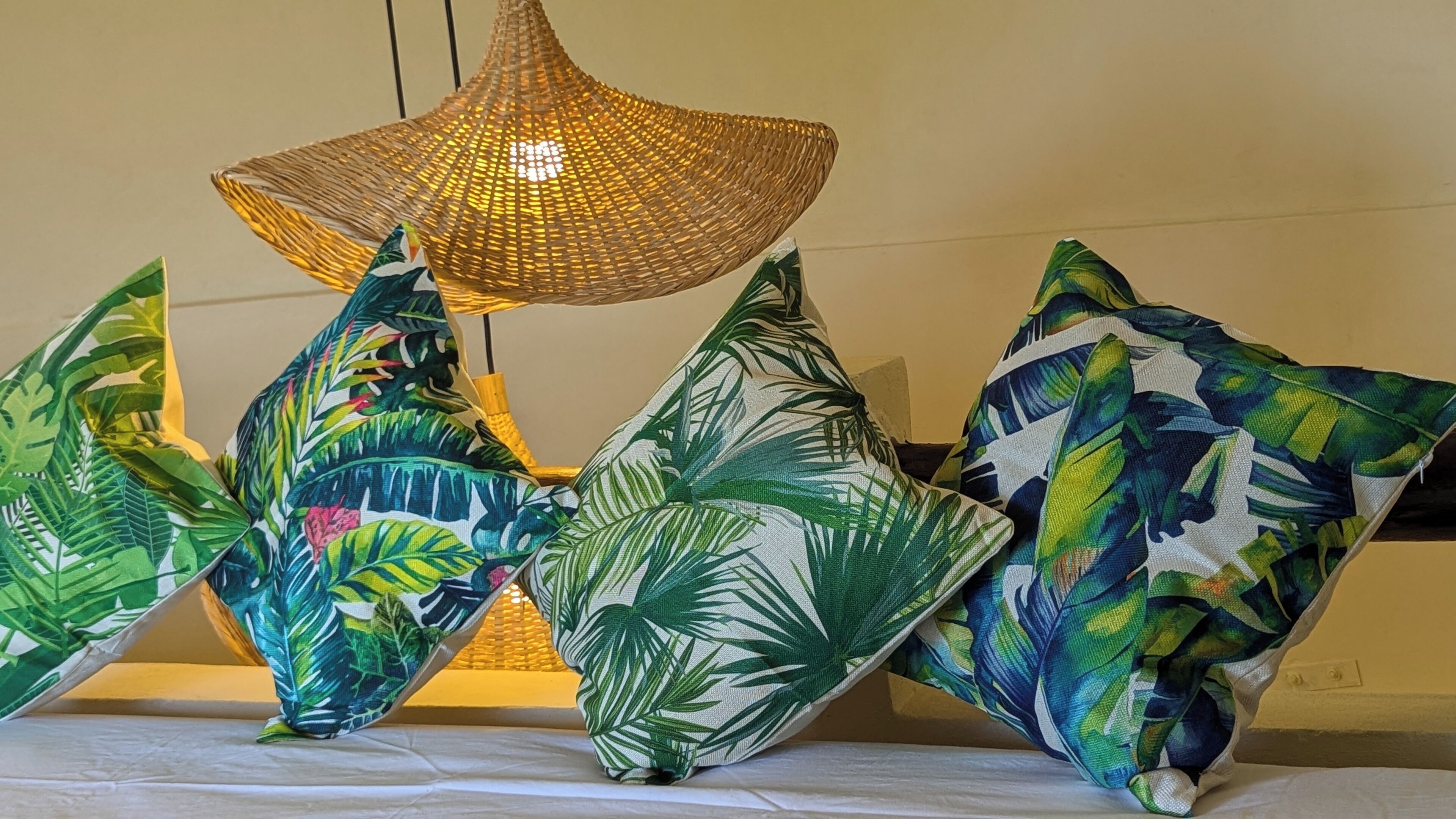 Una cama con sábana blanca, rodeada de almohadones con estampado de hojas tropicales.