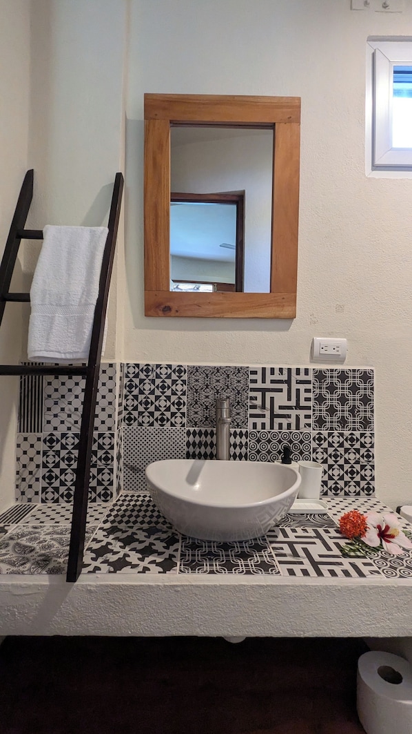 Un baño con un lavamanos blanco, baldosas estampadas en blanco y negro, un toallero de madera, un espejo y un dispensador de papel higiénico.