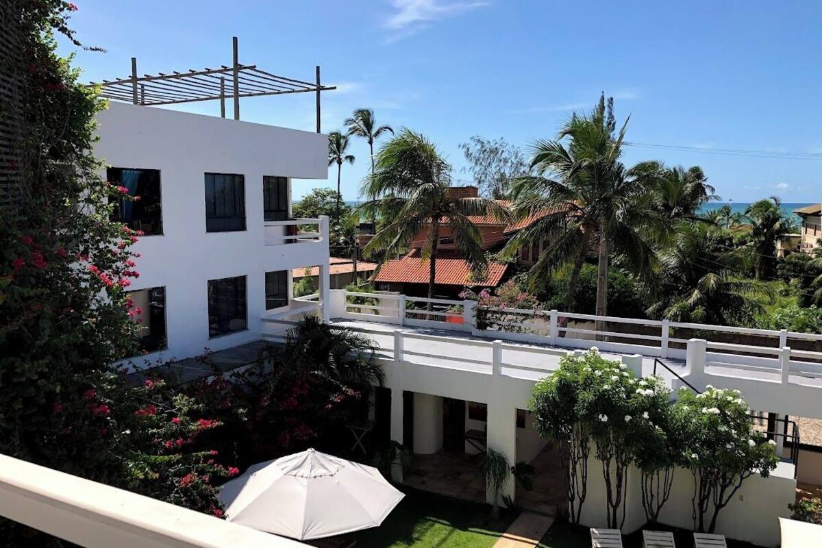 Un bâtiment moderne à l’extérieur blanc, doté d’un espace extérieur couvert avec un parasol blanc, et offrant une vue sur des palmiers et une plage en arrière-plan.