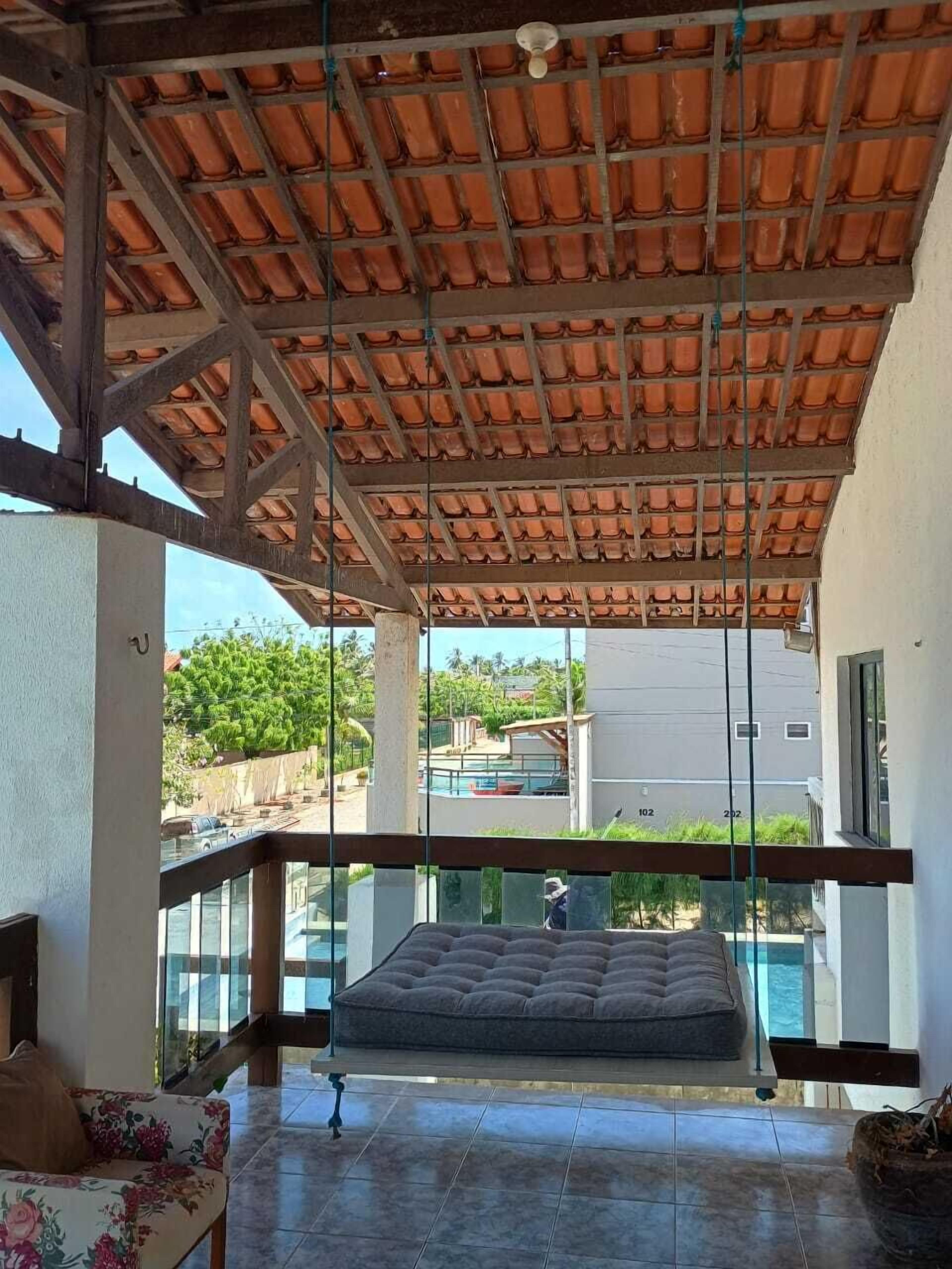 A spacious balcony with a tiled ceiling, a cushioned seating area, and a view of a swimming pool and buildings in the background.