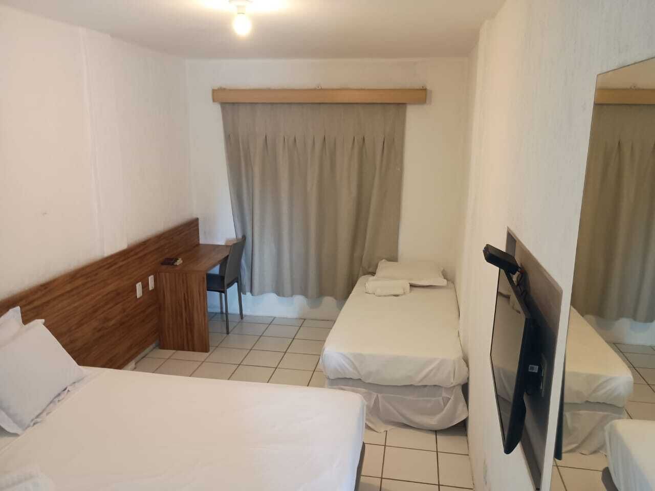 Une chambre d’hôtel avec deux lits simples, un bureau en bois, une chaise, une télévision et une fenêtre avec des rideaux.