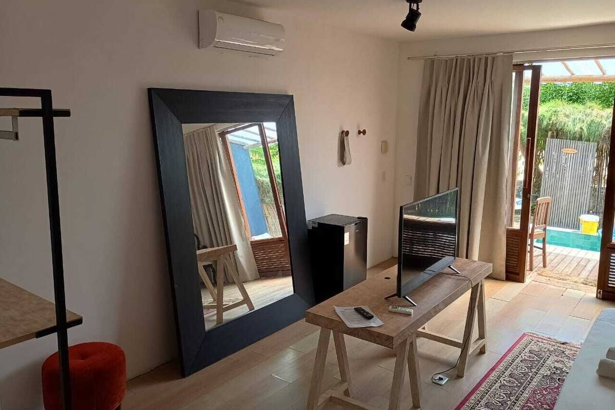 Une pièce dotée d’un grand miroir, d’un téléviseur posé sur une table en bois, d’un fauteuil rouge et d’une vue sur un jardin, accessible par une porte coulissante.
