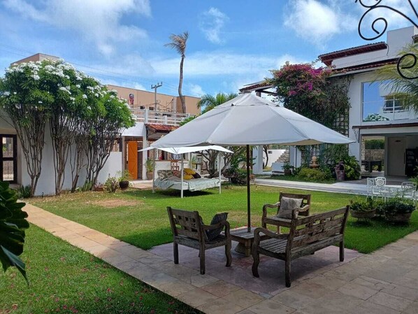 A patio with two wooden benches and a white umbrella, surrounded by a well-maintained lawn and a building with a white facade.