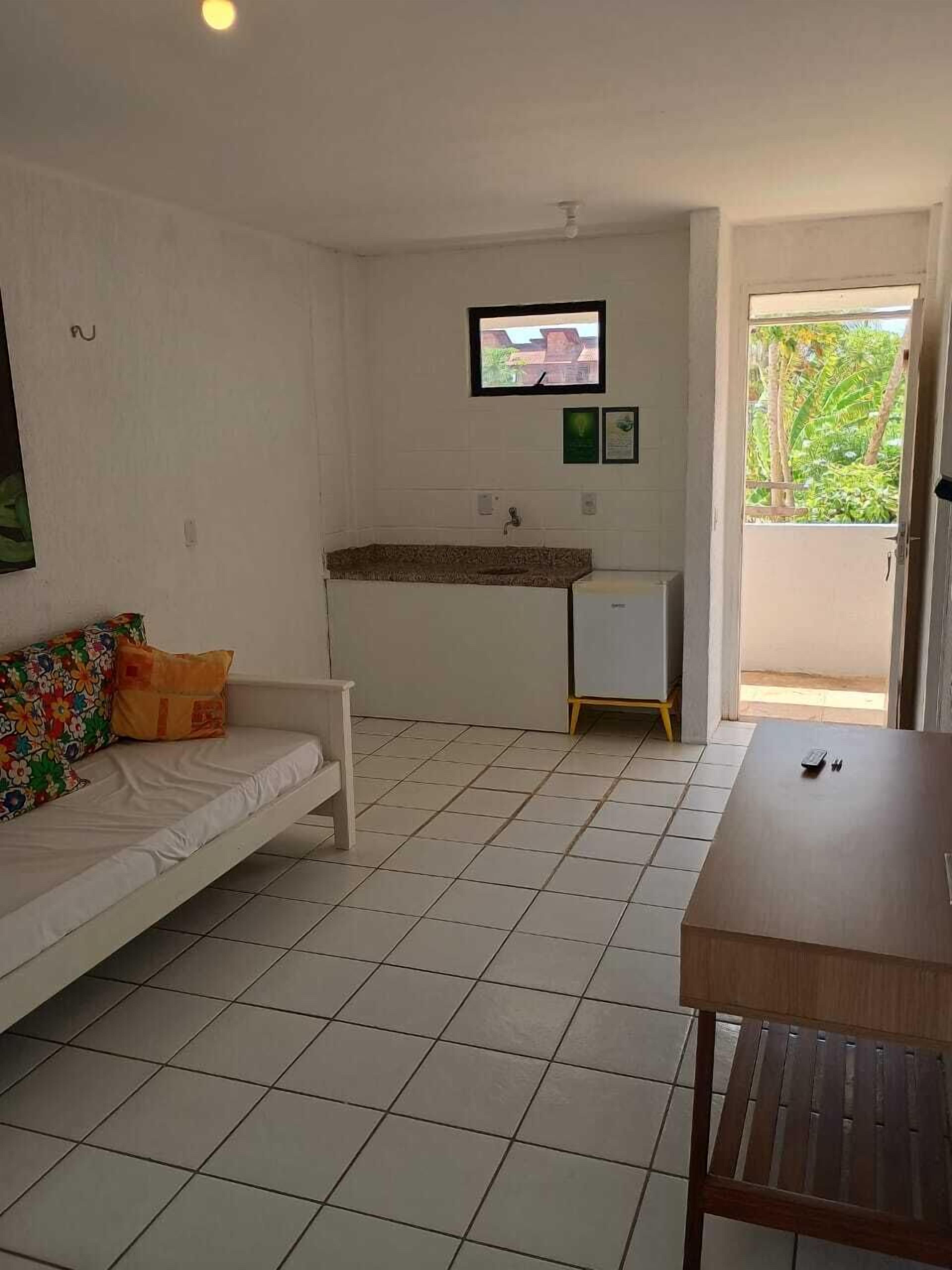 Une pièce avec un canapé blanc, une petite table en bois, un sol carrelé, un coin cuisine et un téléviseur fixé au mur.