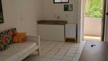 Une pièce avec un canapé blanc, une petite table en bois, un sol carrelé, un coin cuisine et un téléviseur fixé au mur.