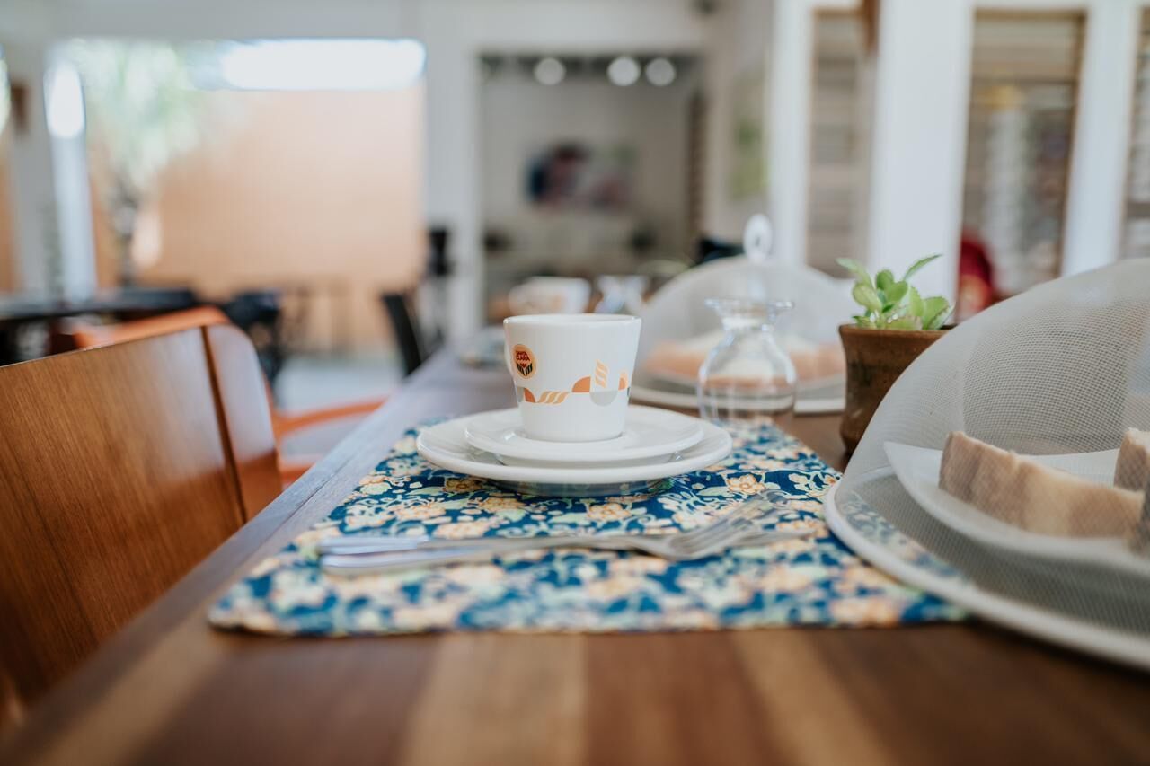 Una mesa de comedor con un mantel individual floral, una taza blanca con un logotipo, una jarra de vidrio y un plato con comida.