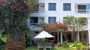 A hotel with a white exterior, a covered outdoor seating area with a white umbrella, and lush greenery including flowering plants. - Inn Tribus (Trairi)