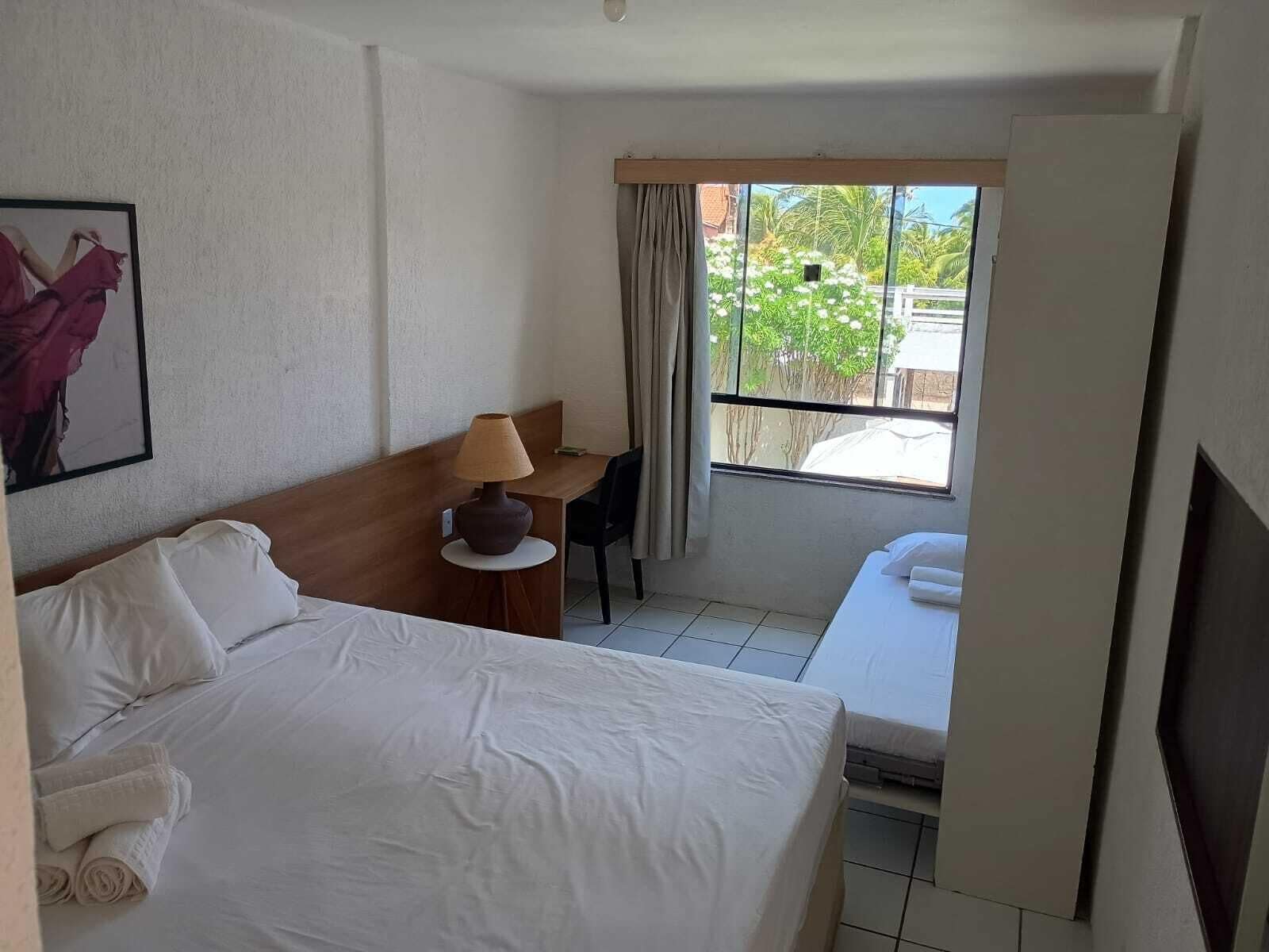 A hotel room with a bed, a desk with a lamp, a chair, and a view of greenery outside.