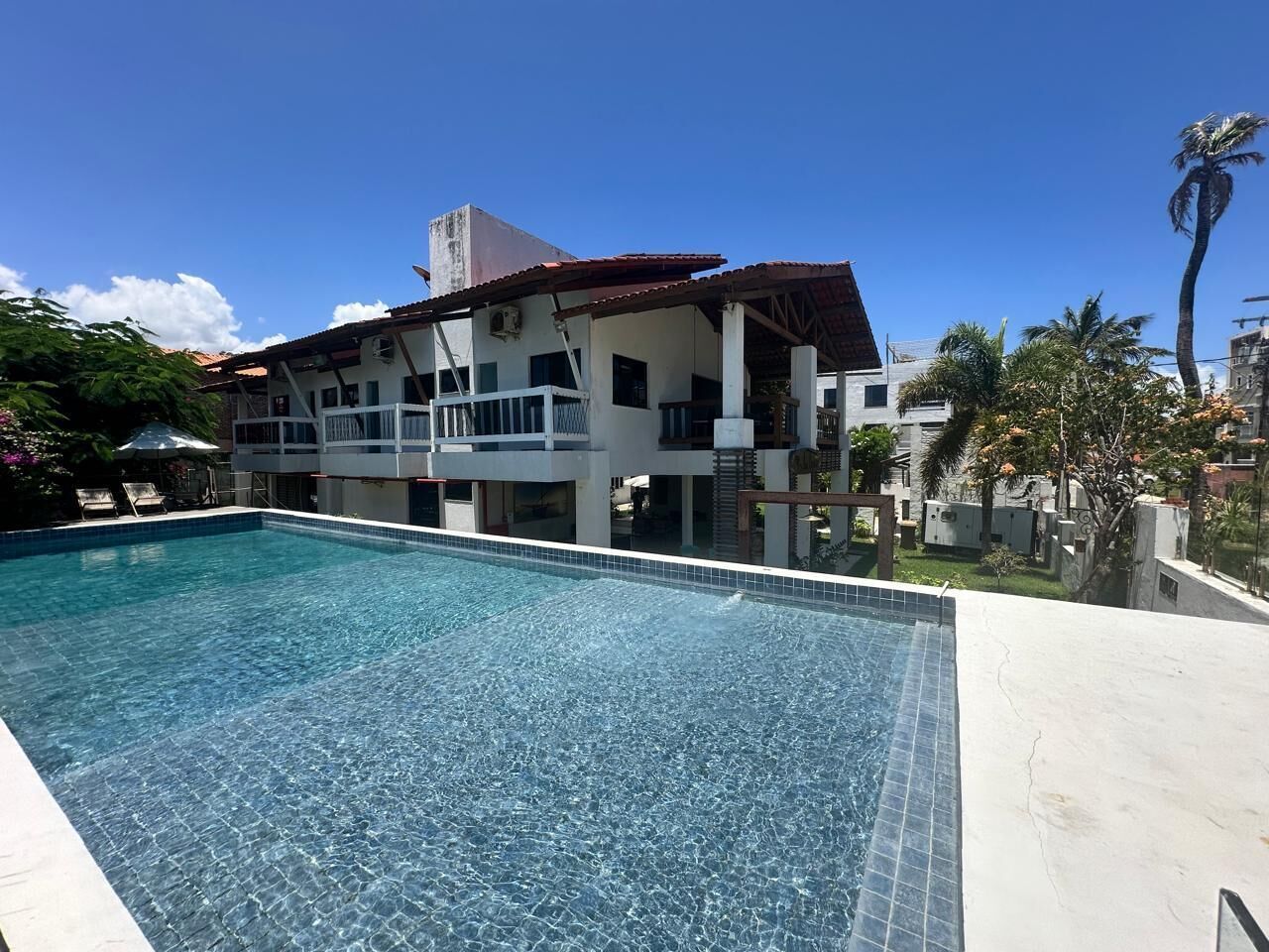 A swimming pool with a tiled edge, a multi-story building with balconies, and palm trees in the background.