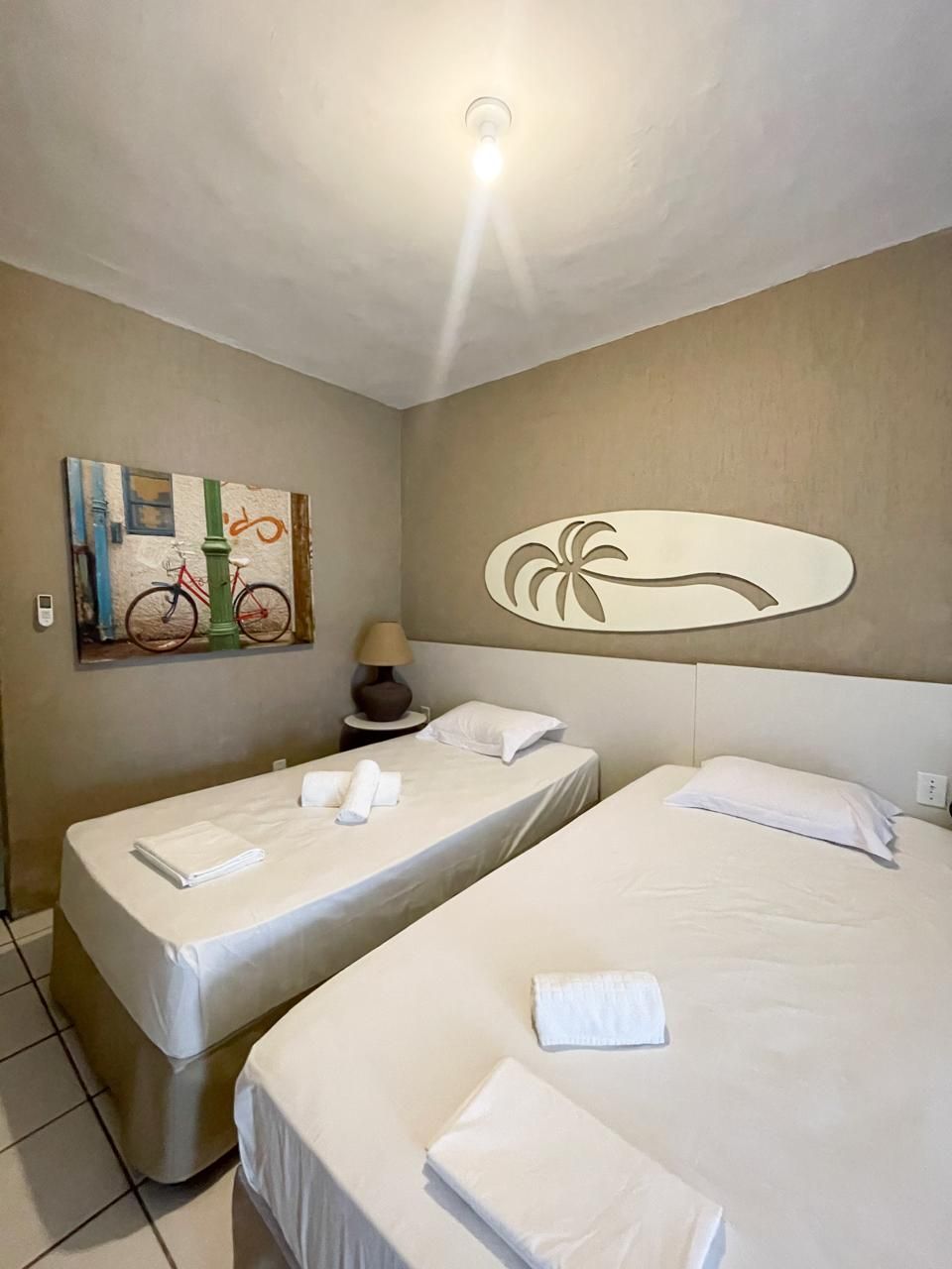 A hotel room with two beds, a painting of bicycles, a palm tree design on the wall, and a ceiling light.