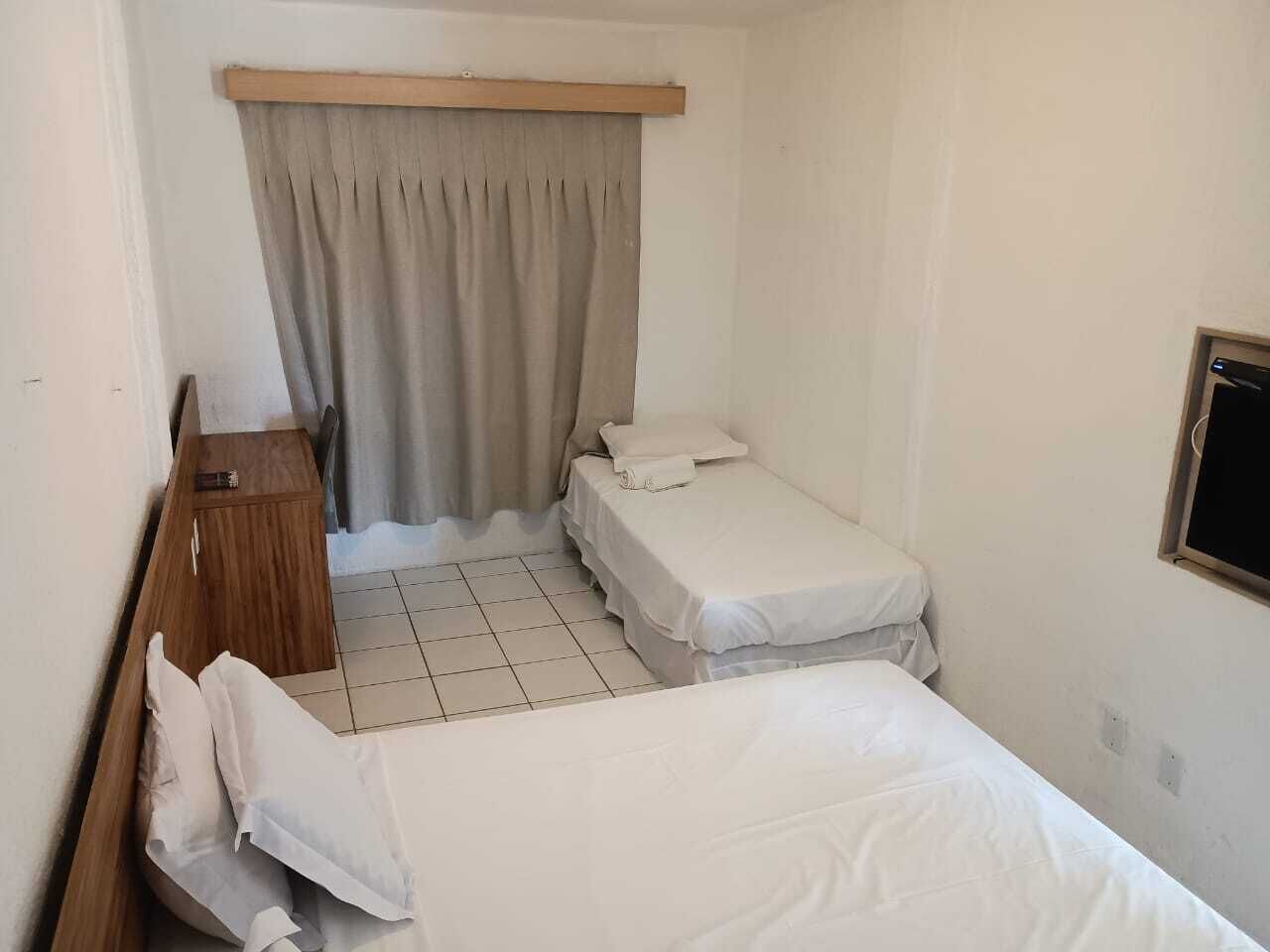 Ein kleines, sauberes Zimmer mit zwei Betten, einem Holznachttisch und einem Fenster mit Vorhängen.