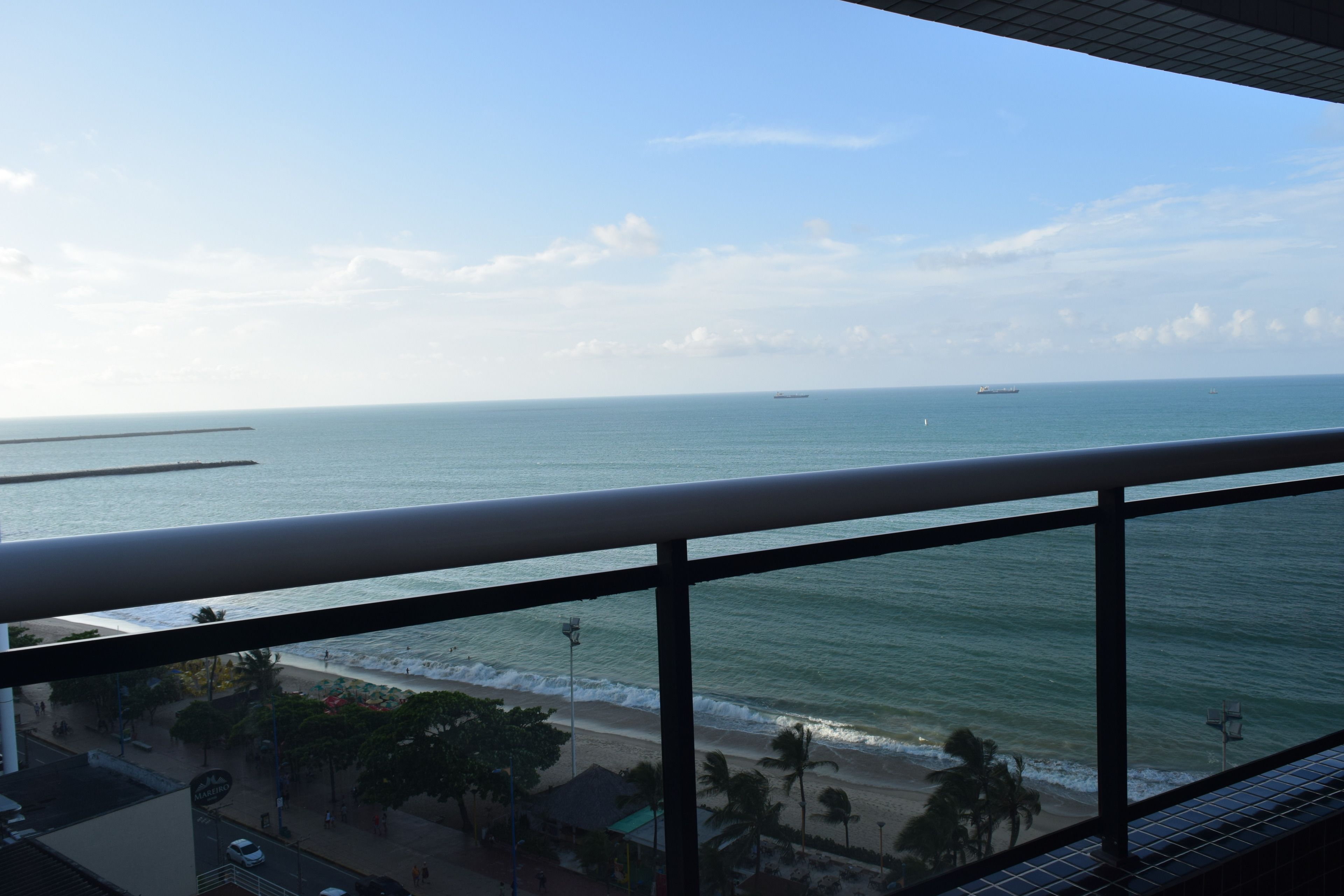A view from a balcony overlooking a beach with a clear sky, a pier, and a few boats in the distance.