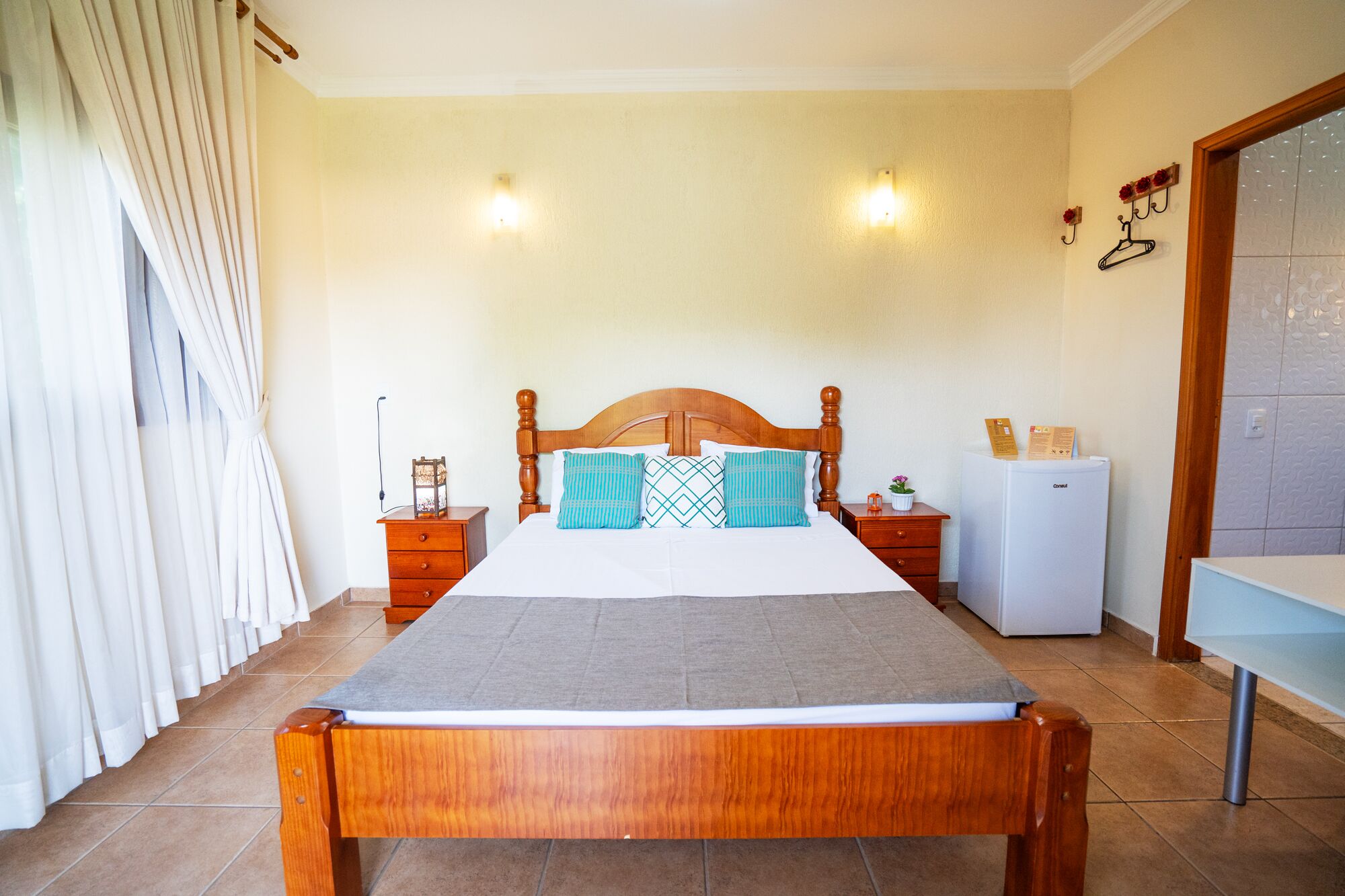 Une chambre à coucher avec un lit en bois, une table de chevet, une commode, un réfrigérateur et une fenêtre avec des rideaux.