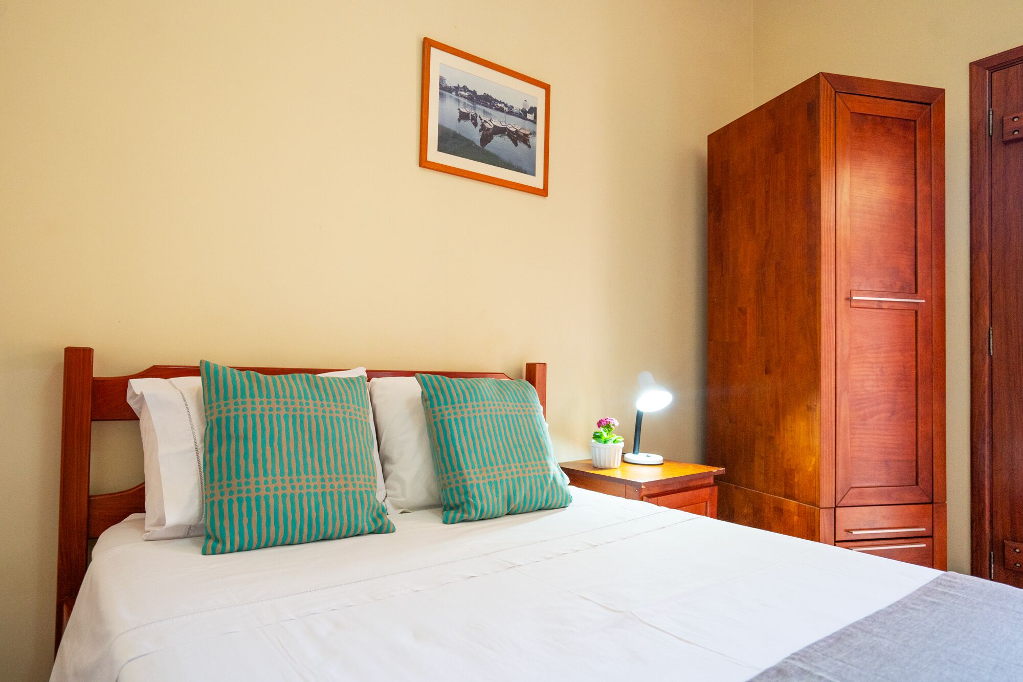 Habitación de hotel con cama, dos almohadas, mesita de noche con lámpara, armario de madera y un cuadro en la pared.