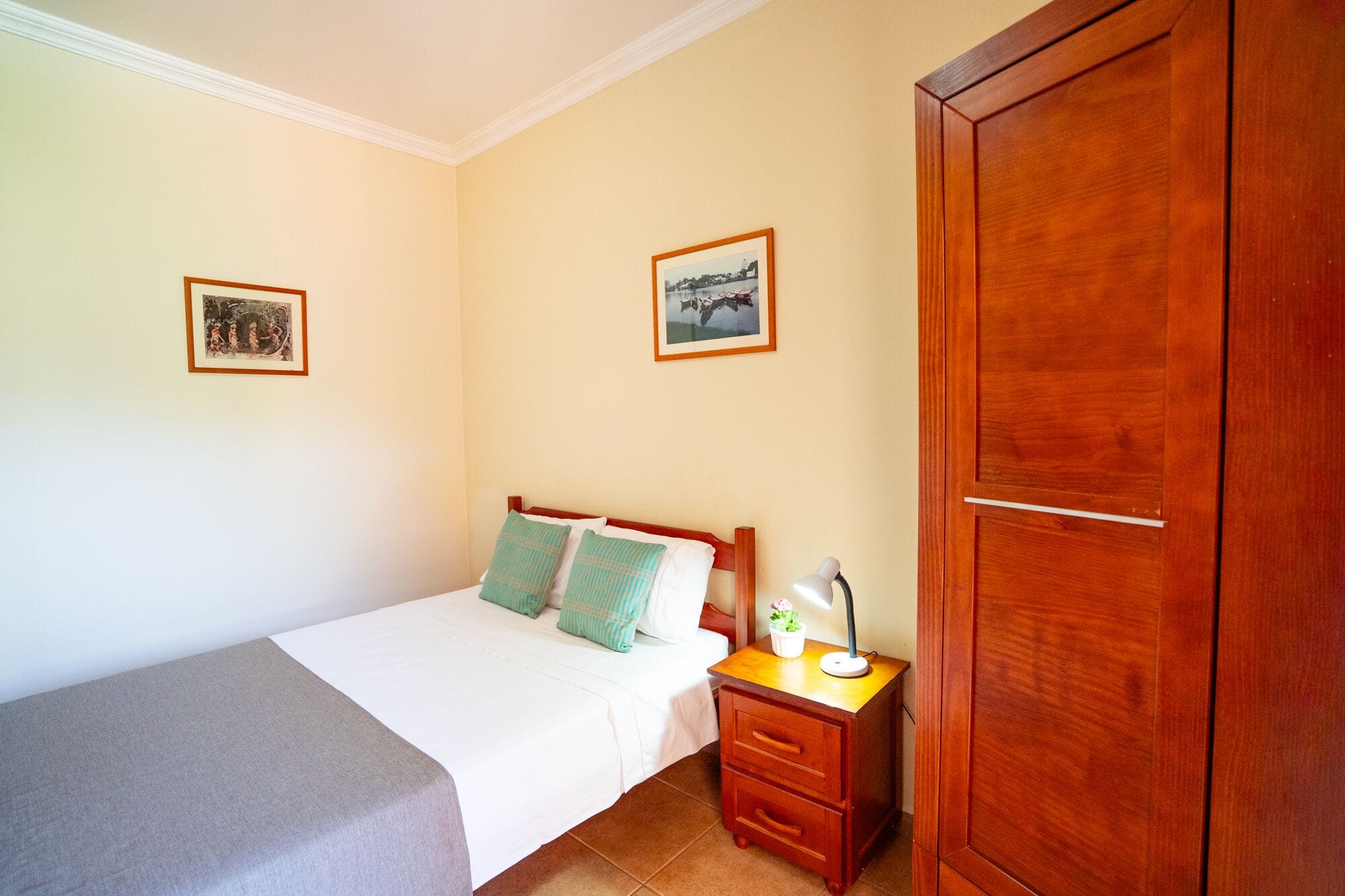 Un dormitorio con cama, mesita de noche, lámpara, armario de madera y dos cuadros enmarcados en la pared.