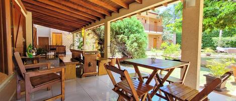 An outdoor patio with wooden furniture, a tiled floor, and a view of the surrounding greenery and buildings.