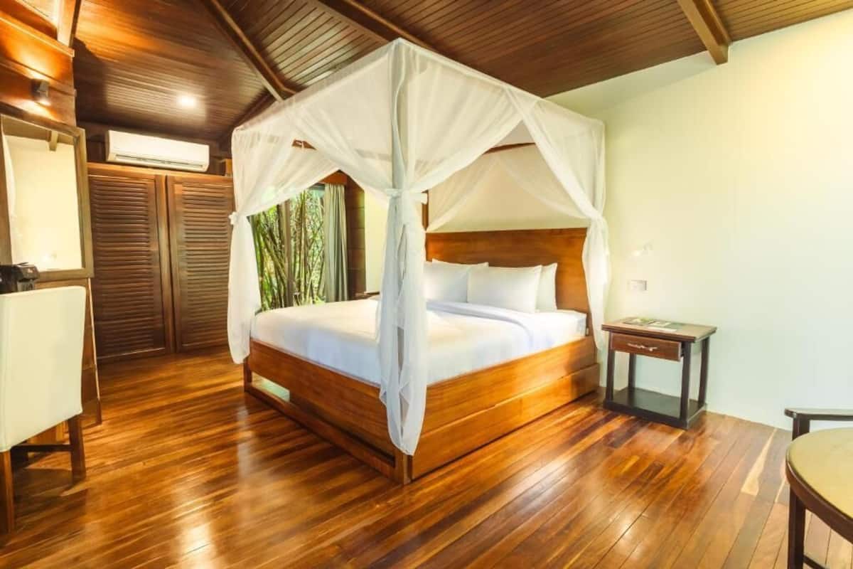 A wooden four-poster bed with a canopy, a bedside table, and a chair in a room with wooden flooring and a sloped ceiling.