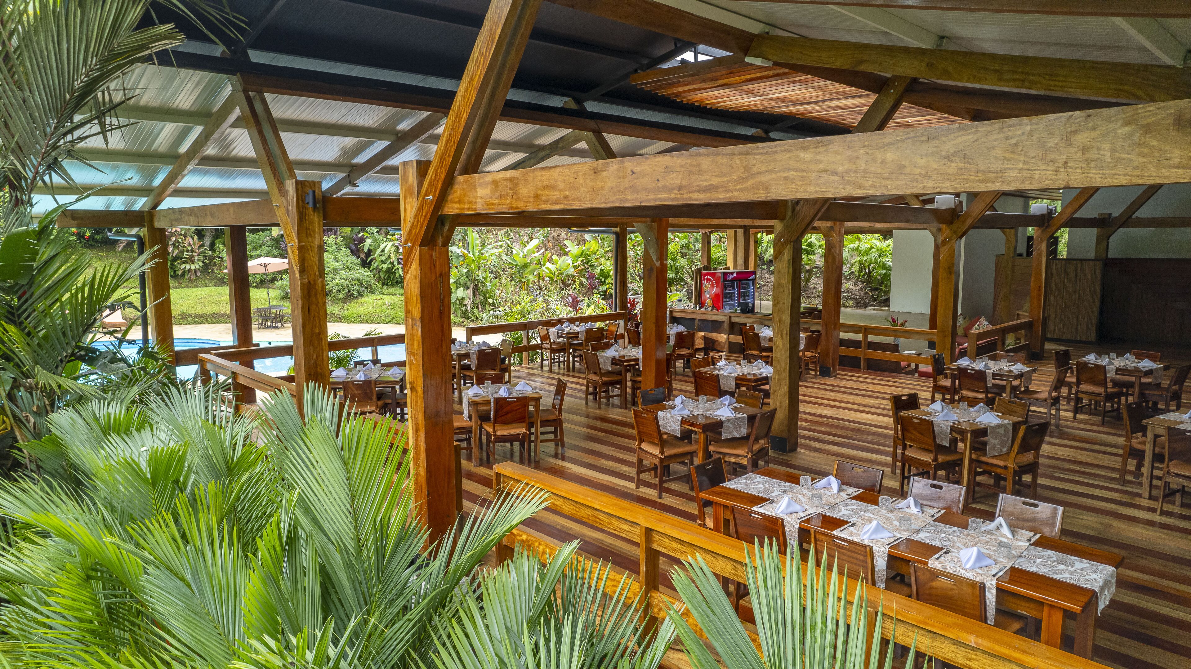 A covered outdoor dining area with wooden tables and chairs, surrounded by lush greenery and a pool in the background.