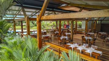 A covered outdoor dining area with wooden tables and chairs, surrounded by lush greenery and a pool in the background.