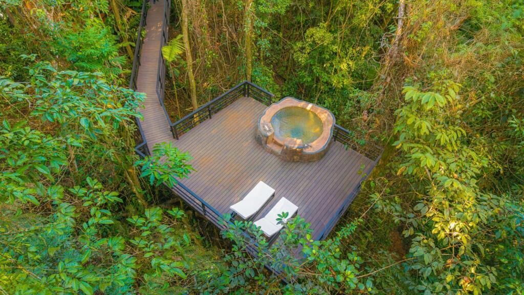 A hot tub surrounded by lush greenery, a wooden deck, and two lounge chairs.