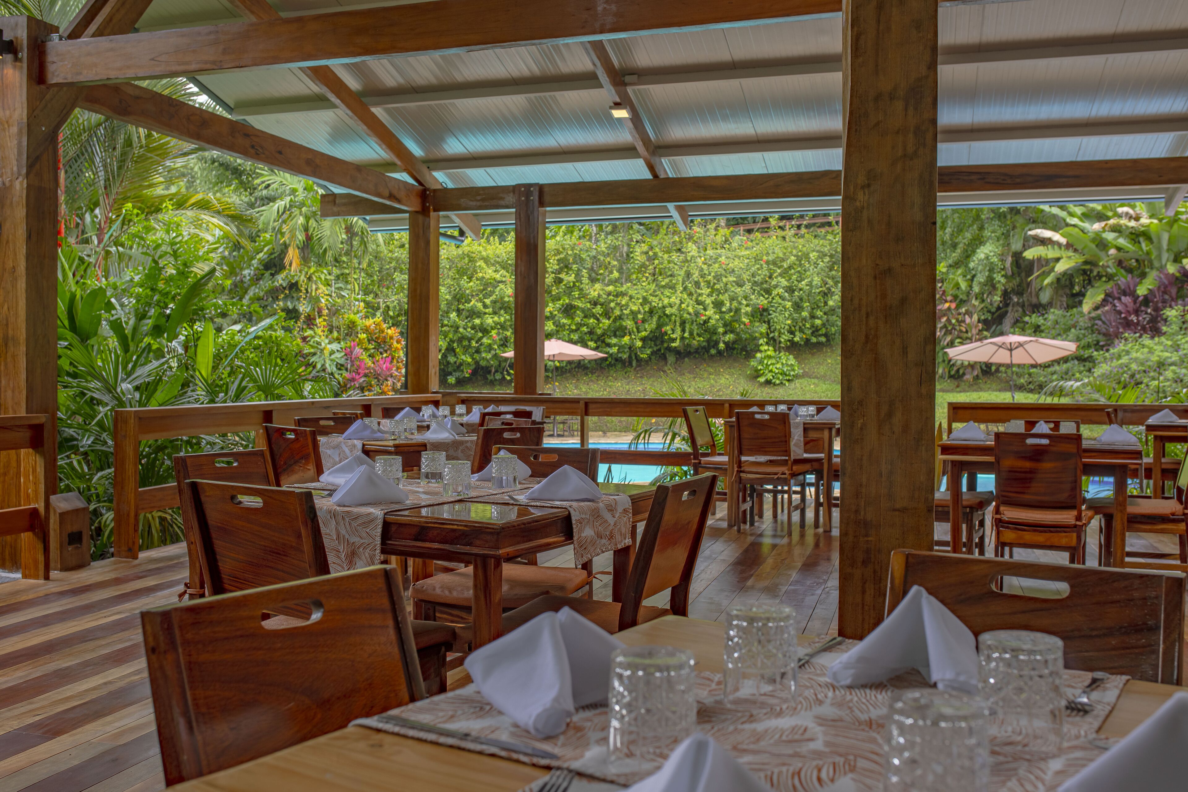 A restaurant with wooden tables and chairs, a covered patio, and a view of a lush garden and pool.
