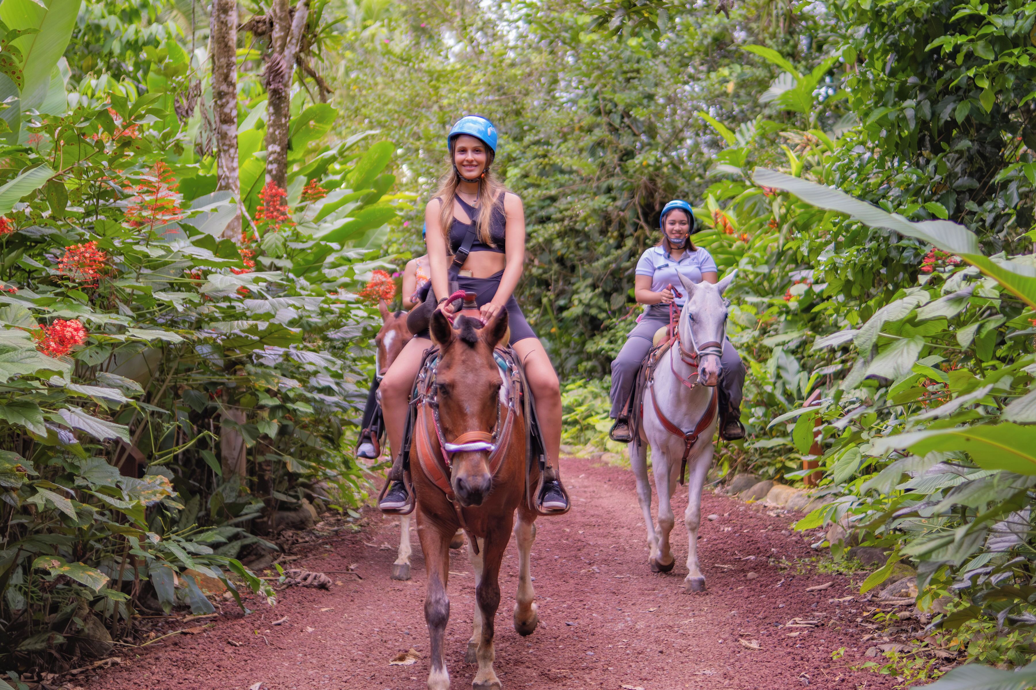 Dos personas montando a caballo por un sendero de tierra rodeado de abundante vegetación.