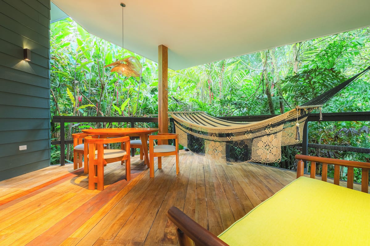 A wooden deck with a hammock, chairs, and a table, surrounded by lush tropical foliage.