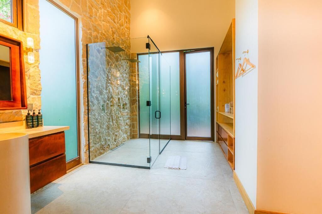 A bathroom with a stone wall, a glass shower enclosure, a wooden vanity with a mirror, and a shelf with toiletries.