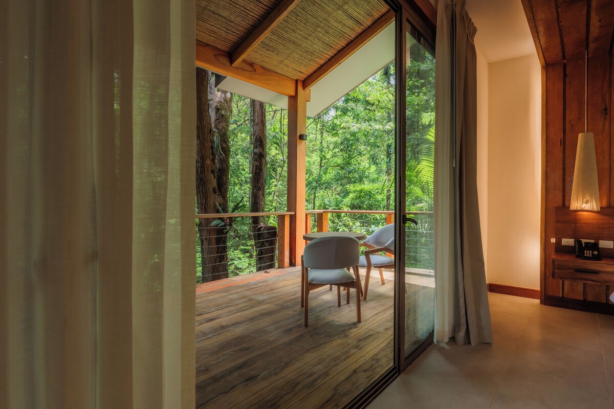 A bedroom with a large window, wooden flooring, a bed, a nightstand, and a chair.