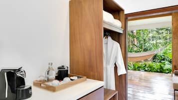 A hotel room with a wooden vanity, a coffee machine, a tray with toiletries, and a view of a hammock and greenery outside.