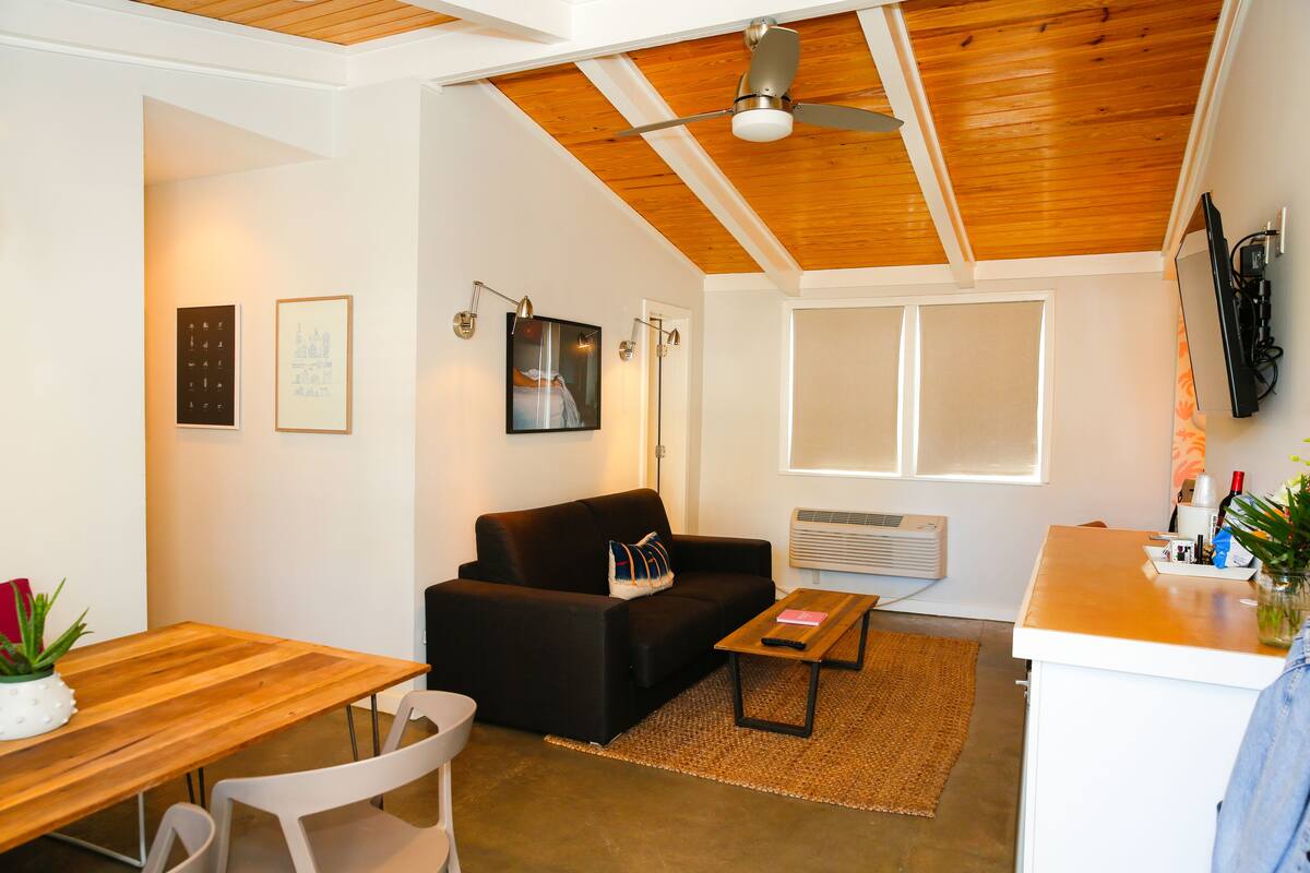 A modern living room with a wooden ceiling, a black sofa, a wooden dining table, and a white wall with framed pictures.