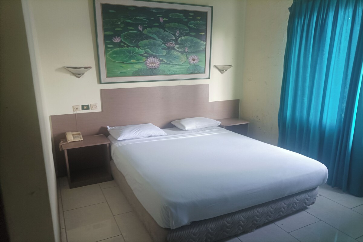 Quarto de hotel com cama, mesa de cabeceira com telefone, quadro de nenúfares e cortina azul.