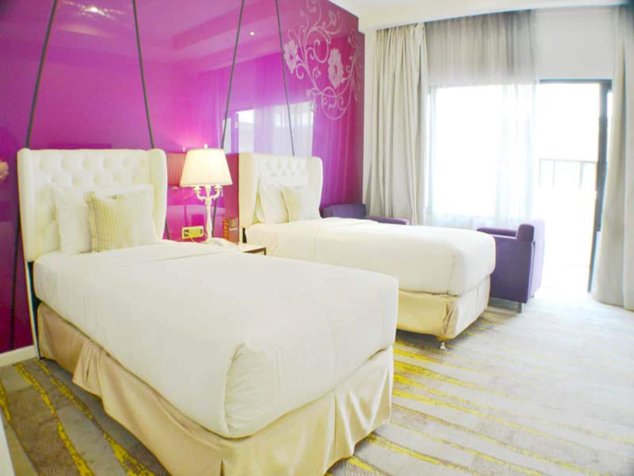 A hotel room with two beds, a large window with curtains, and a bright pink wall.