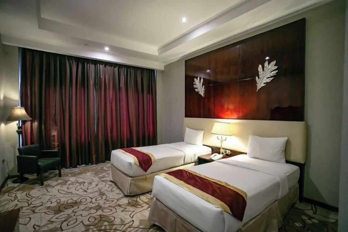 A hotel room with two beds, a large window with curtains, a desk with a chair, and a decorative wall piece.