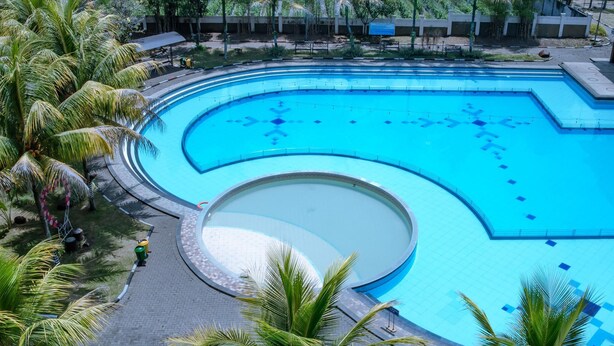 A large, curved swimming pool surrounded by palm trees and a paved walkway. - Sutan Raja Hotel and Convention Cen (Soreang)