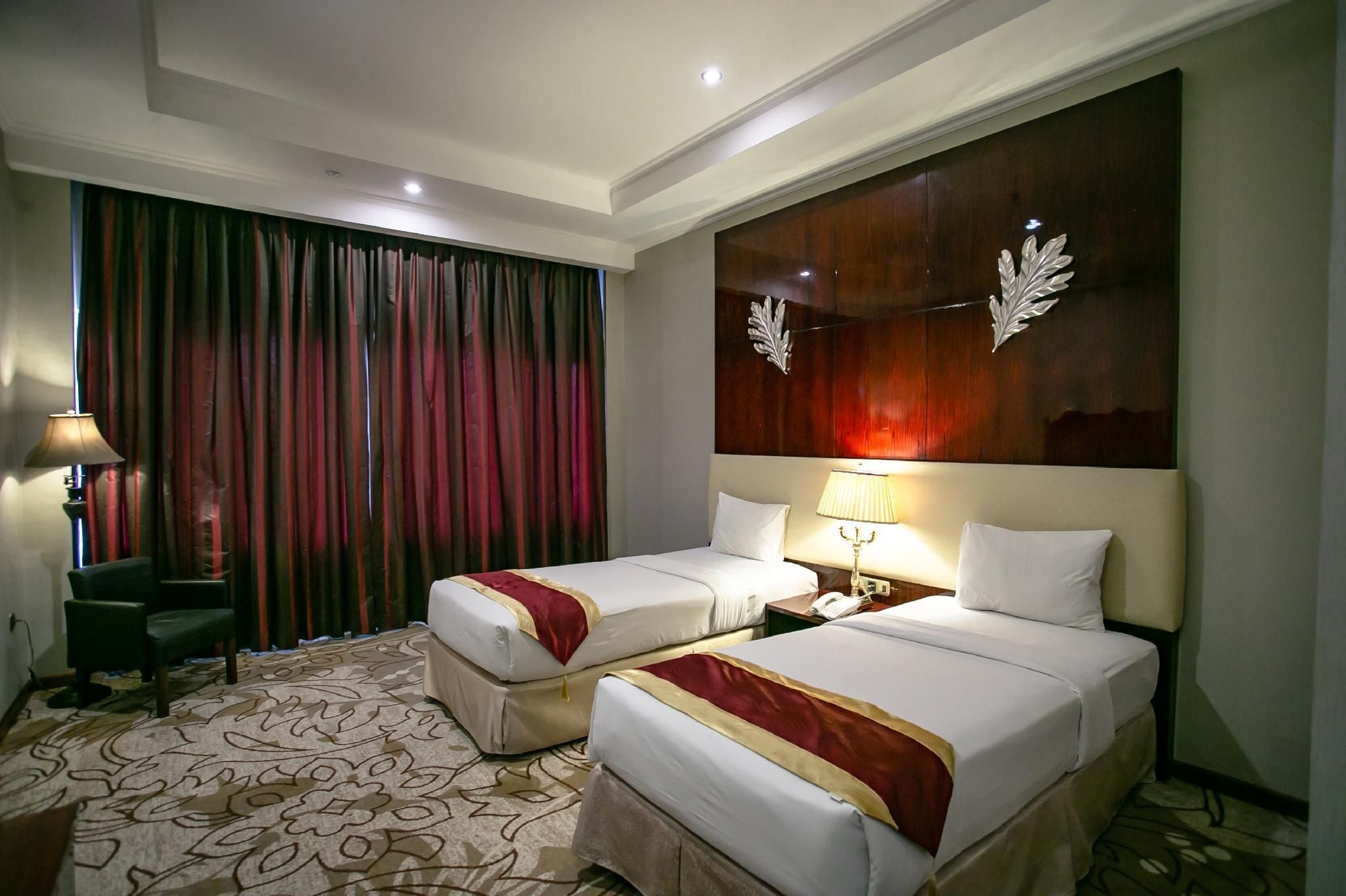 A hotel room with two beds, a large window with curtains, a desk with a lamp, and a decorative wall panel.