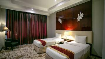 Quarto de hotel com duas camas, uma janela grande com cortinas, uma mesa com luminária e um painel decorativo na parede.