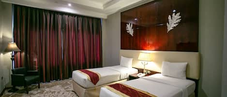 A hotel room with two beds, a large window with curtains, a desk with a lamp, and a decorative wall panel.