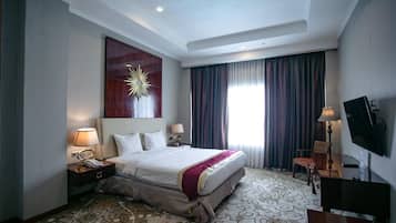 Habitación de hotel con cama, escritorio, silla, televisor, ventana grande con cortinas y un elemento decorativo en la pared.