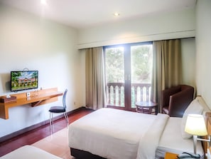 A hotel room with a bed, a desk with a television, a chair, a small table, and a balcony with curtains.
